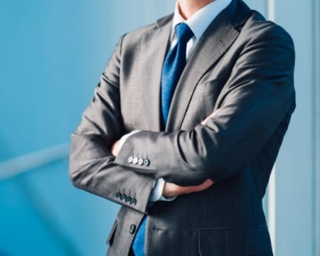 グレーのスーツに青いネクタイを締めた男性が腕を組んで立っている姿

